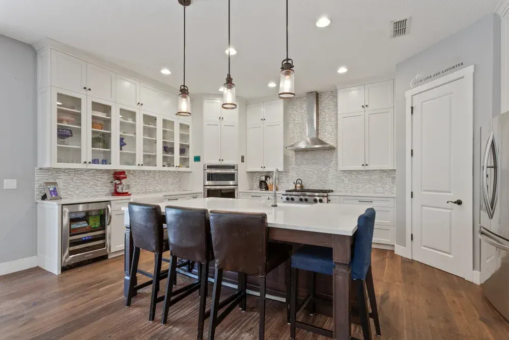 Modern white kitchen with island, bar stools, cabinets, and stainless steel appliances.