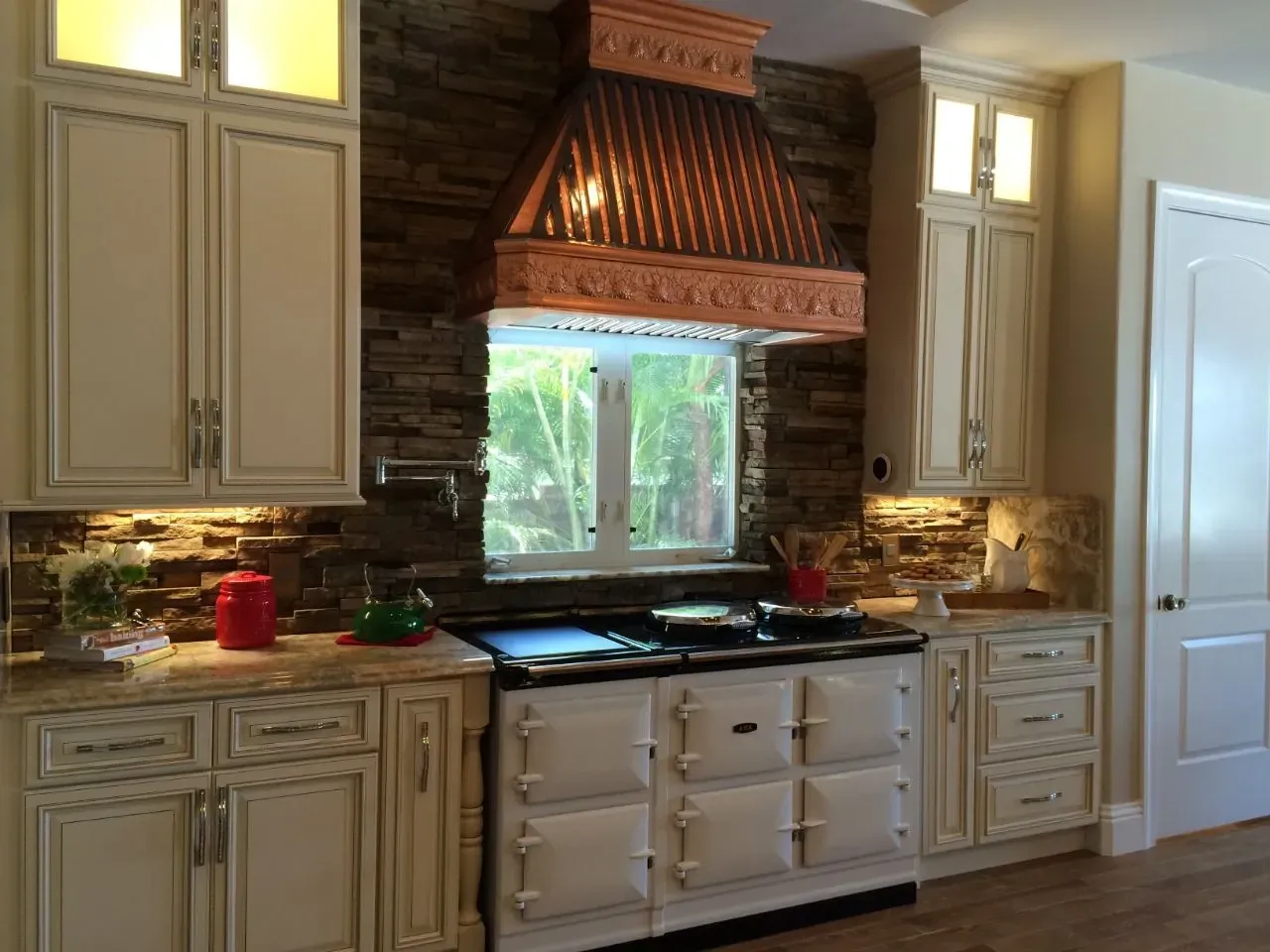Cream-colored kitchen with a white stove, stone backsplash, copper range hood, and cabinets; a window is behind the stove.
