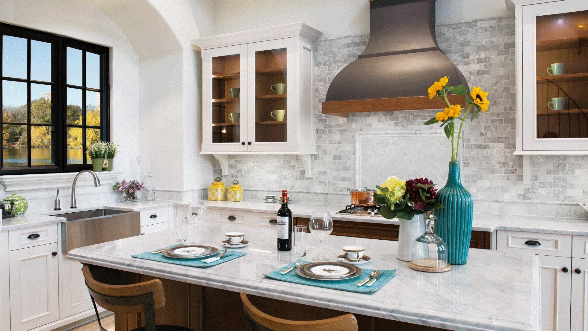 Bright kitchen with island, two place settings, flowers, and stainless steel sink by a window.