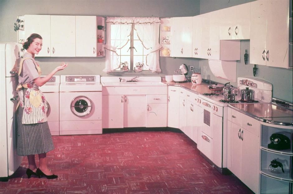 Woman in retro kitchen with white cabinets, appliances, and red flooring. She gestures near the washing machine.