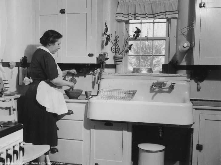 Woman in apron prepares food at a sink in a vintage kitchen, black and white photo.