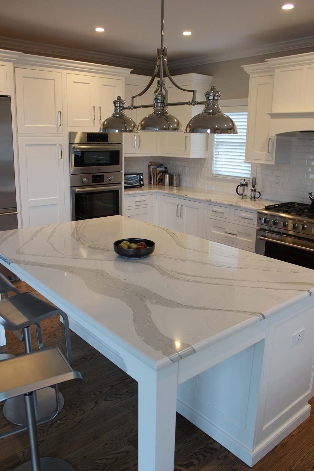 White kitchen with large island and stainless steel appliances. Overhead lighting, white cabinets, and marble countertops.