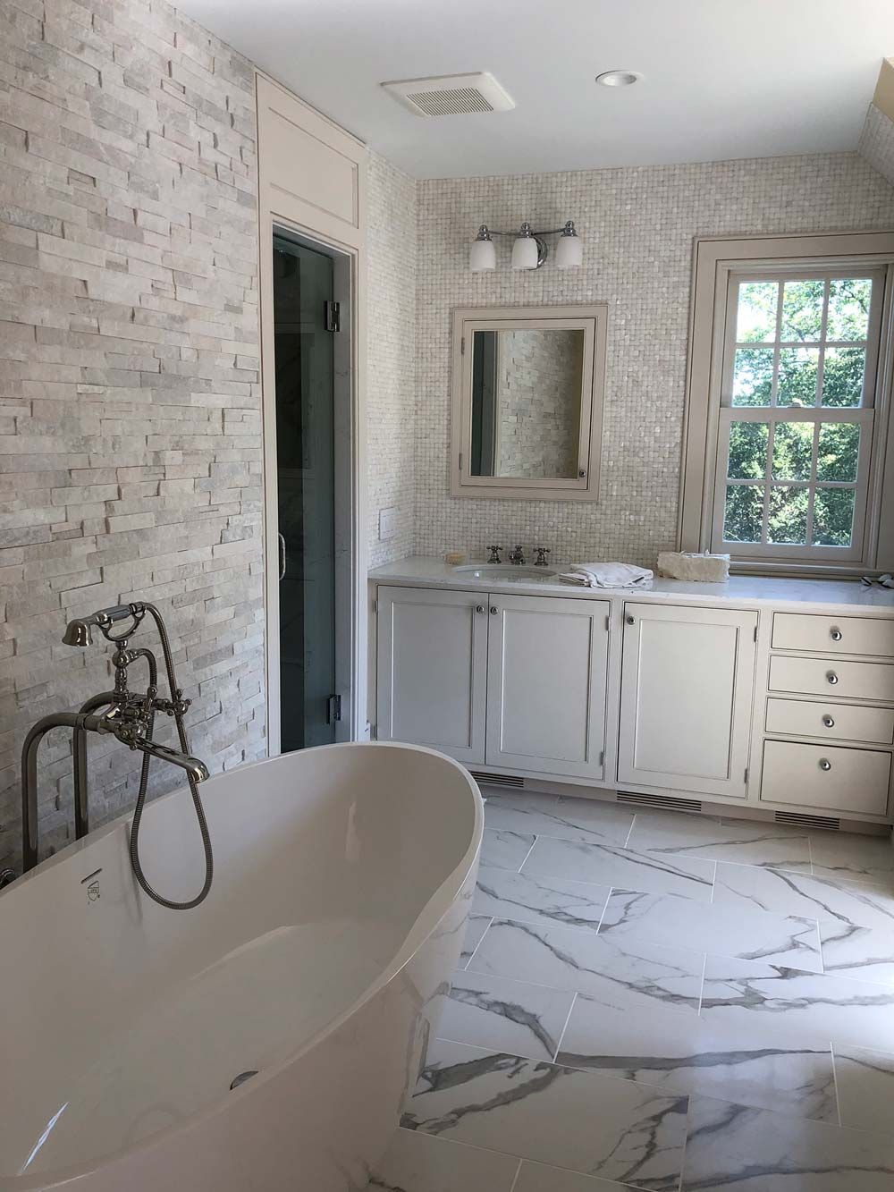 Bathroom with white stone tile, white cabinets, soaking tub, and a window.
