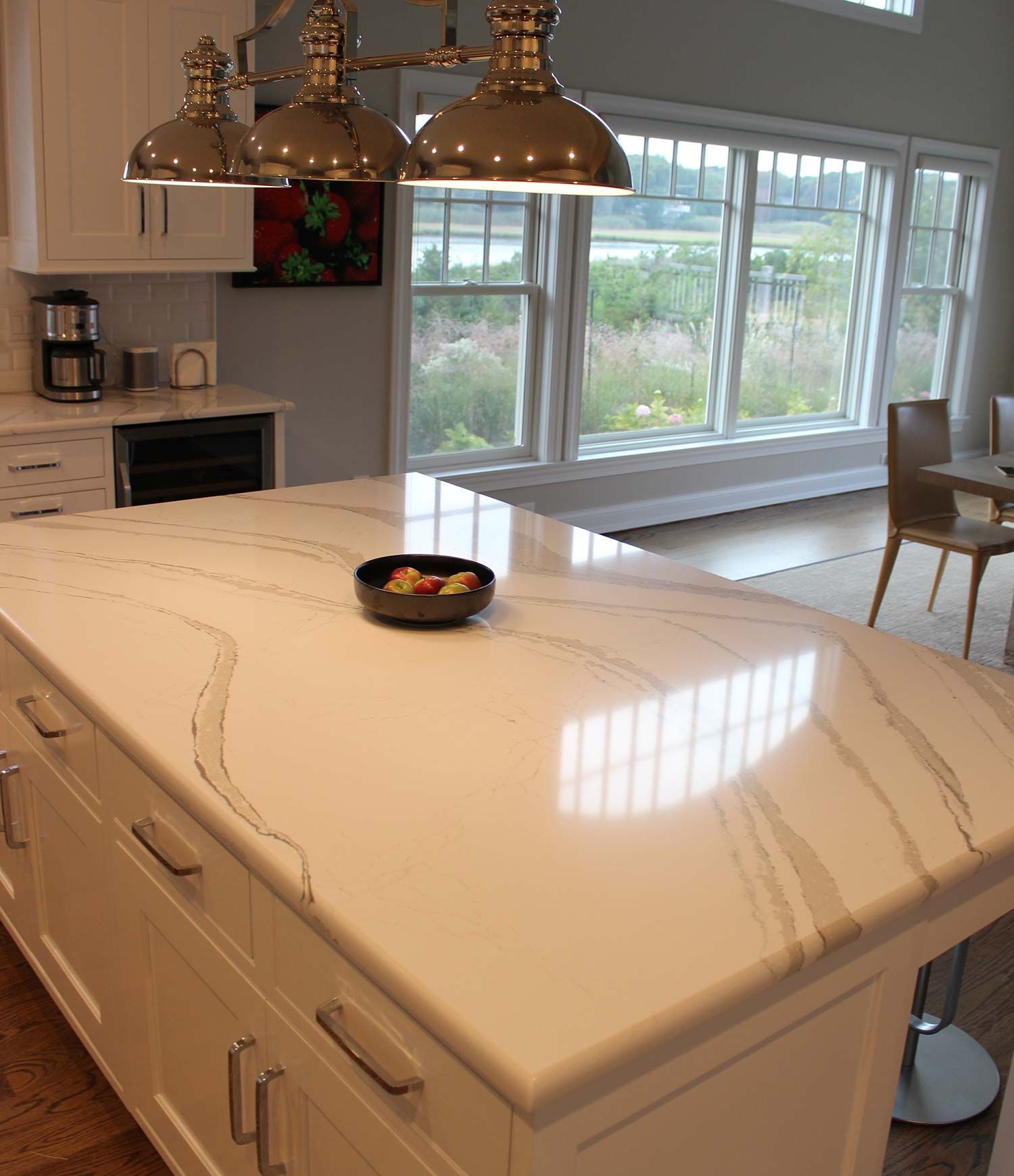 White kitchen island with veined countertop, pendant lights, windows overlooking a landscape.