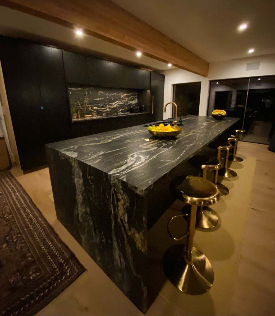 Black granite kitchen island with brass stools and faucet, dark cabinets.