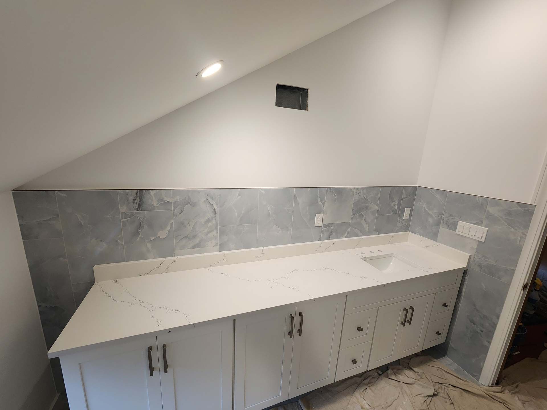 Bathroom vanity with white countertop, gray backsplash, white cabinets, angled wall, and overhead lighting.