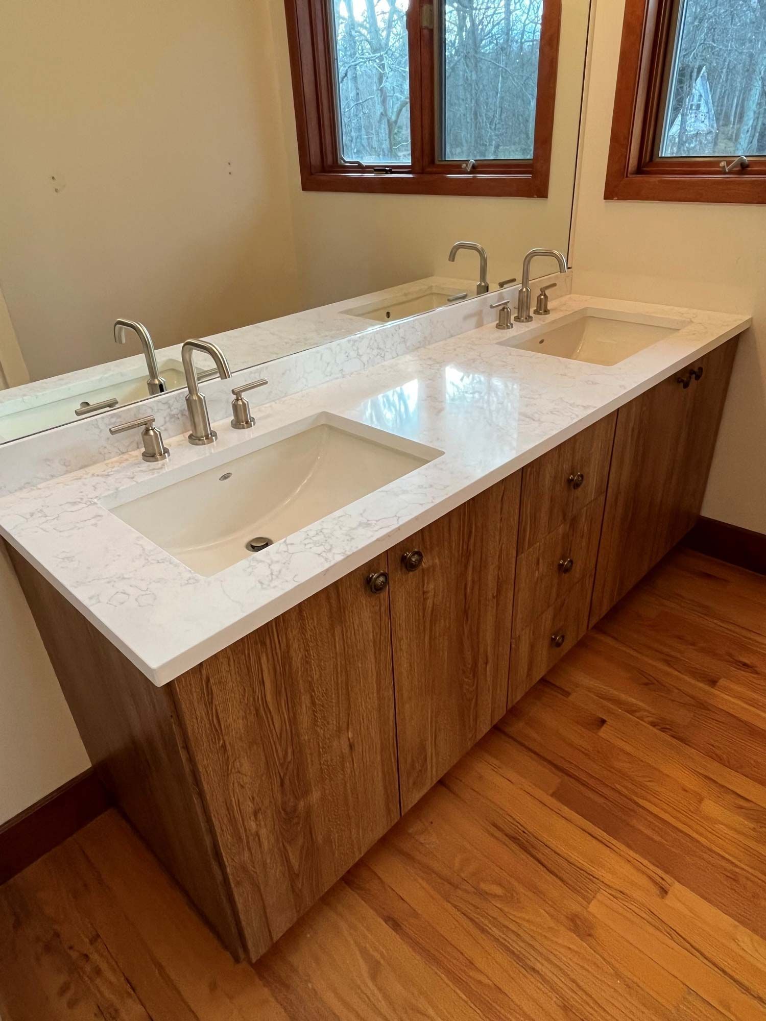 Wooden vanity with two sinks, white countertop, and silver faucets, in a bathroom with wood flooring and a large mirror.