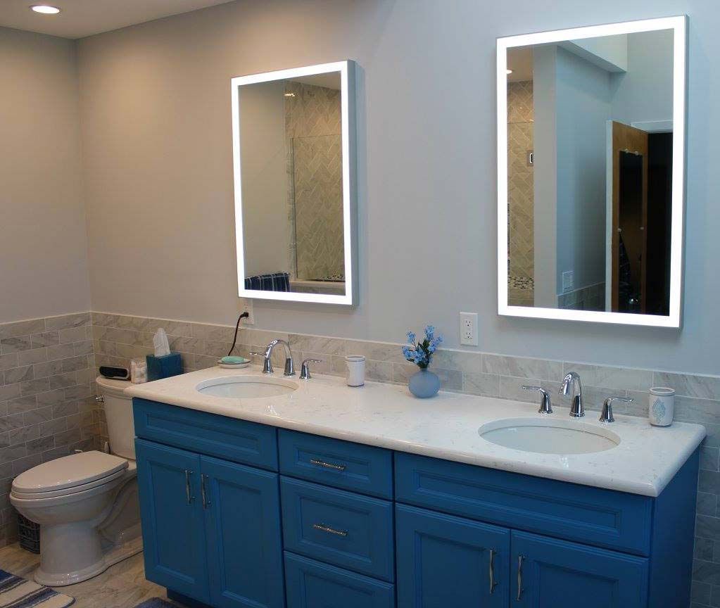 Bathroom with blue vanity, white countertop, two sinks, and lit mirrors.