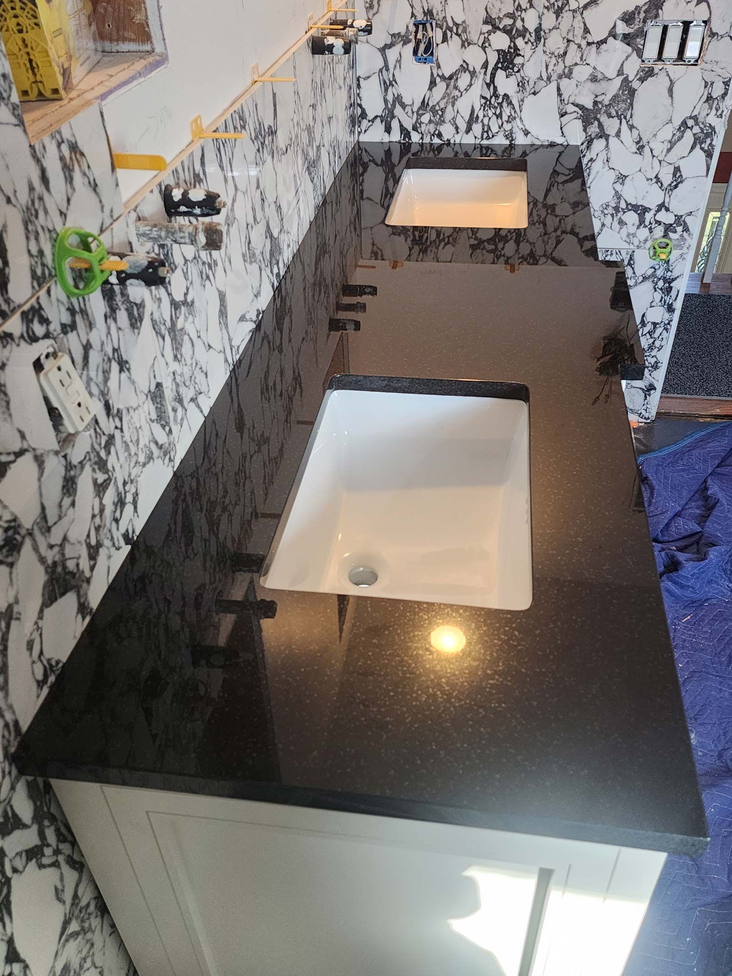 Black granite countertop with two white sinks, against a marble-patterned wall.