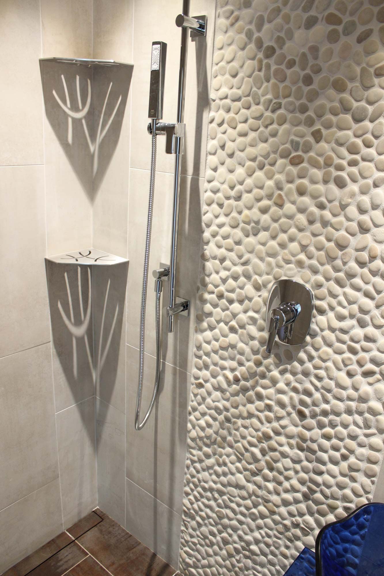 Shower with pebble tile wall and corner shelves, chrome fixtures.