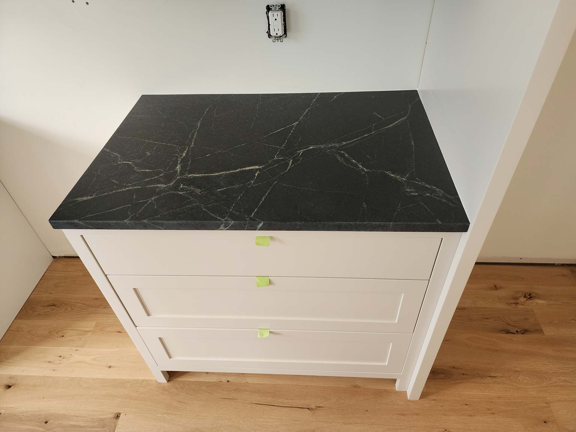 White cabinet with black countertop and veining, three drawers. Located near a wall and wood flooring.