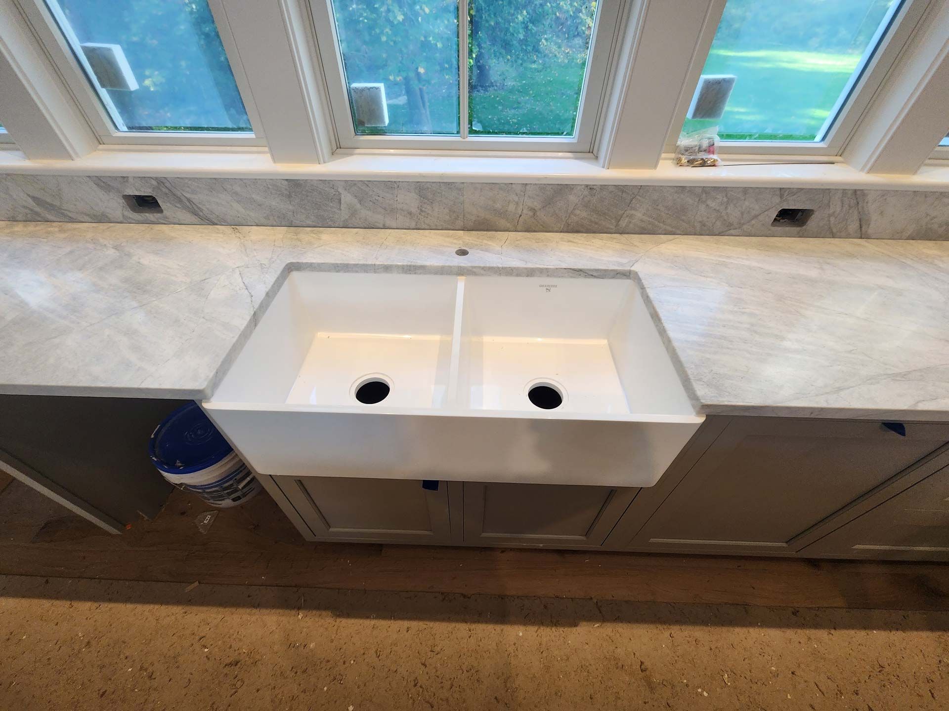 White farmhouse-style kitchen sink in a grey countertop, cabinets below, and windows above.