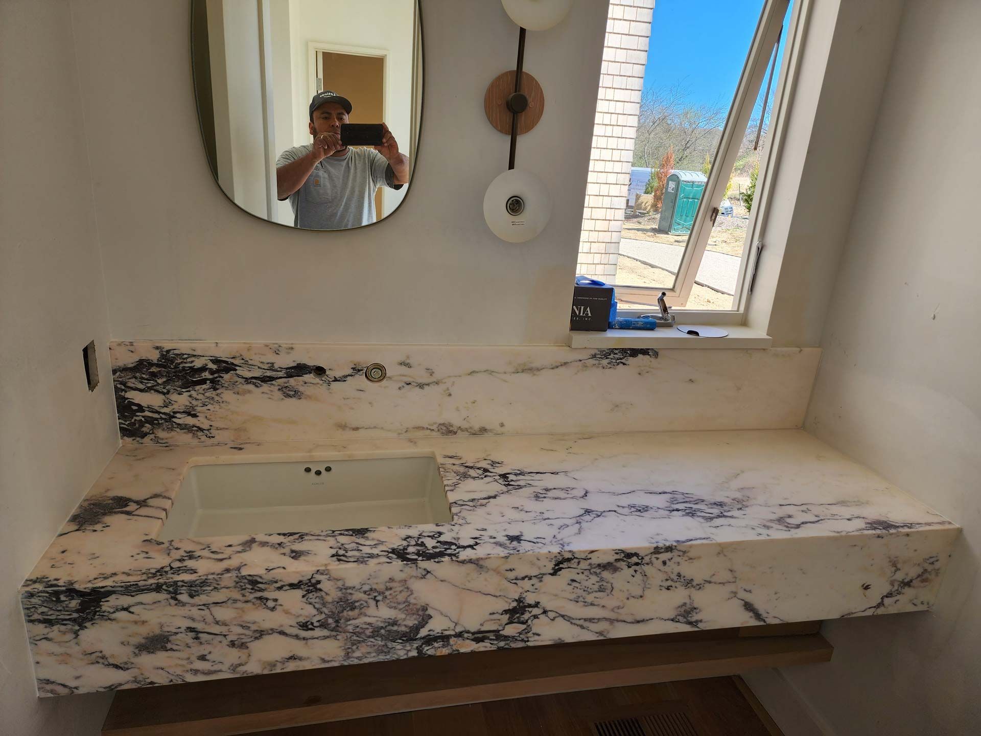 Bathroom vanity with white countertop, sink, and oval mirror; person taking a photo.
