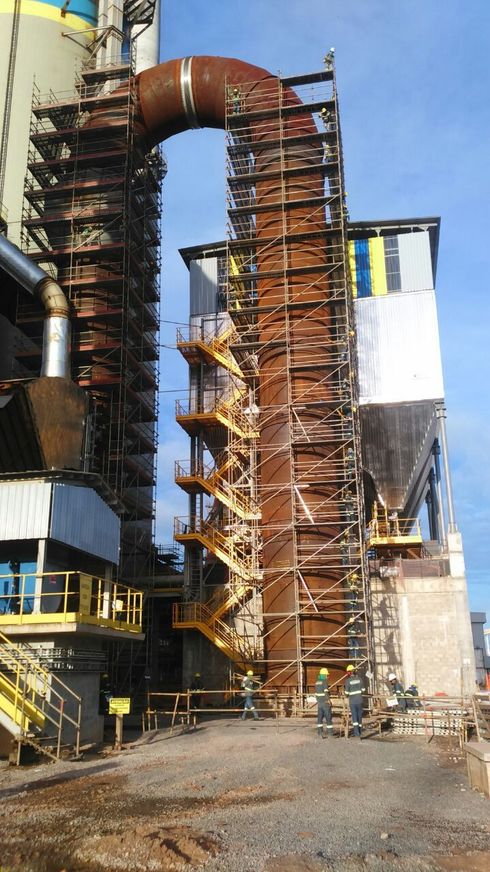 Estrutura industrial alta com andaimes, canos enferrujados e escada externa sob um céu azul.