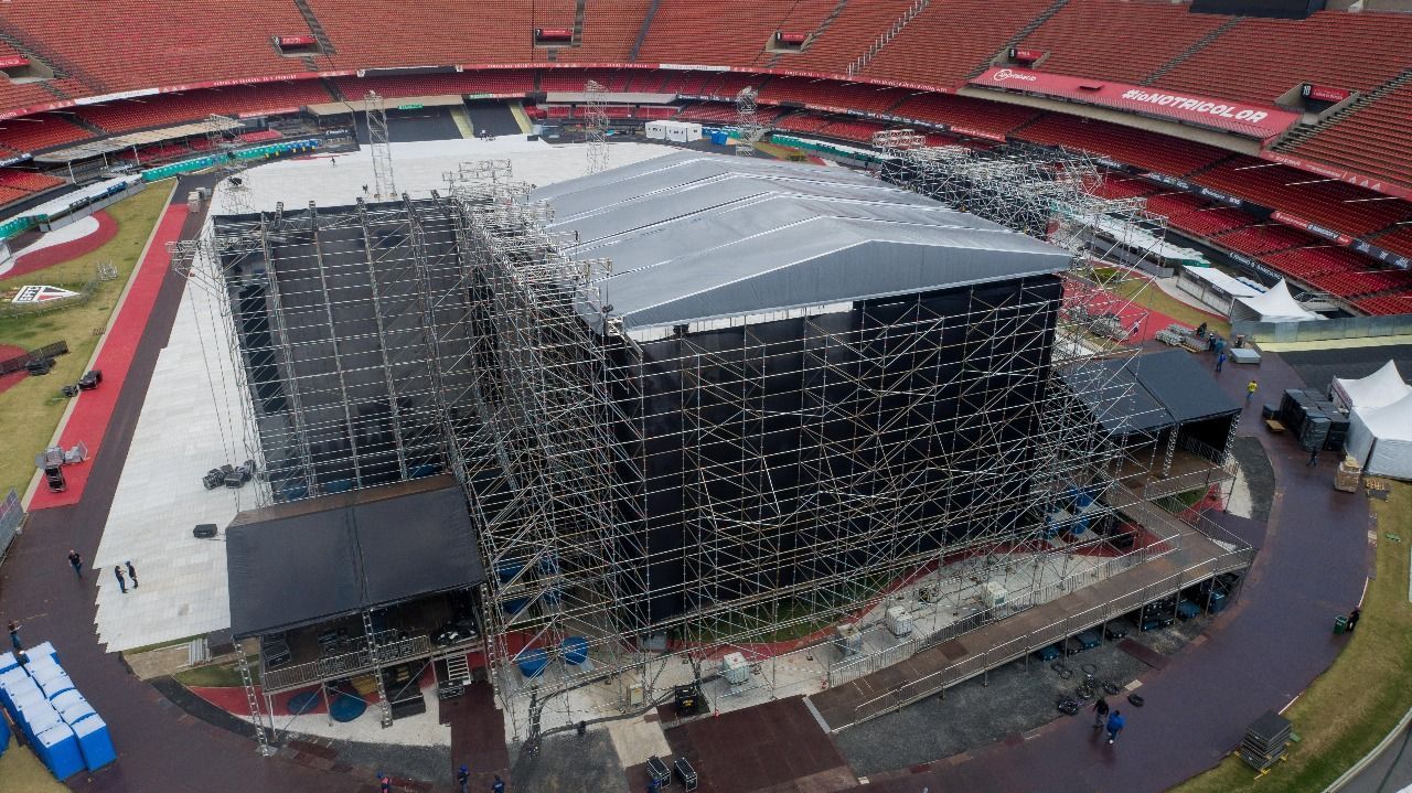 Palco de concerto com assentos pretos e vermelhos na grama; céu do pôr do sol.