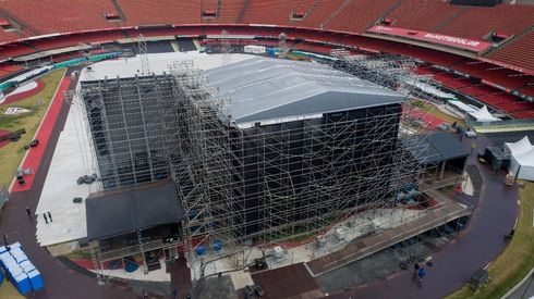 Palco de concerto com assentos pretos e vermelhos na grama; céu do pôr do sol.