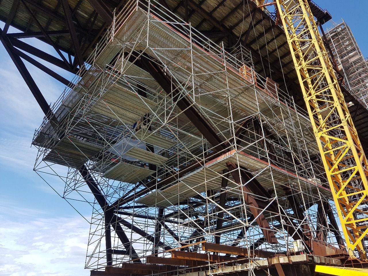 Vista de ângulo baixo de uma ponte em construção com andaimes e vigas de aço contra um céu azul.