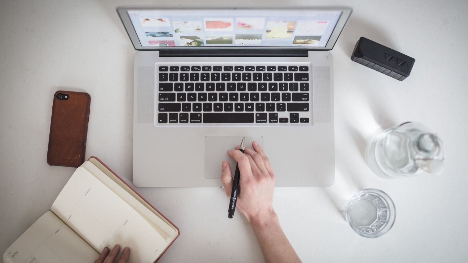 Person working on a laptop at a white desk with a notebook, phone, water, and mouse.