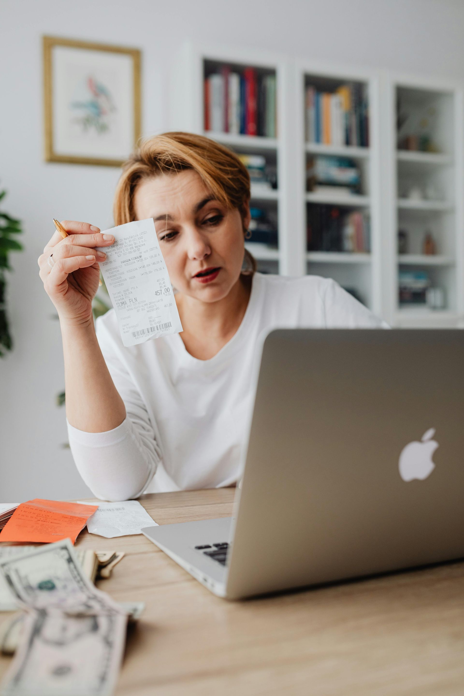 Woman holding receipt, looking at laptop, with money on desk.