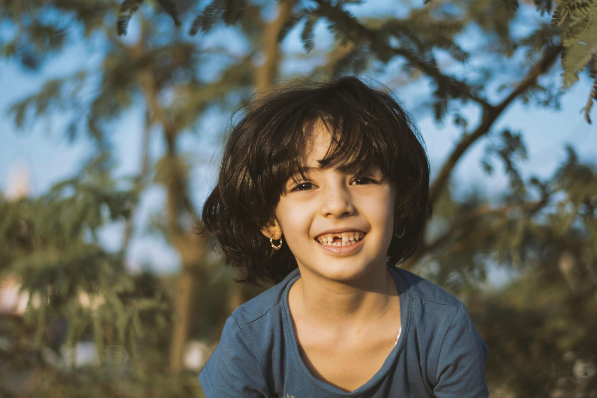 Smiling child with missing front tooth, blue shirt, outdoors near green trees.