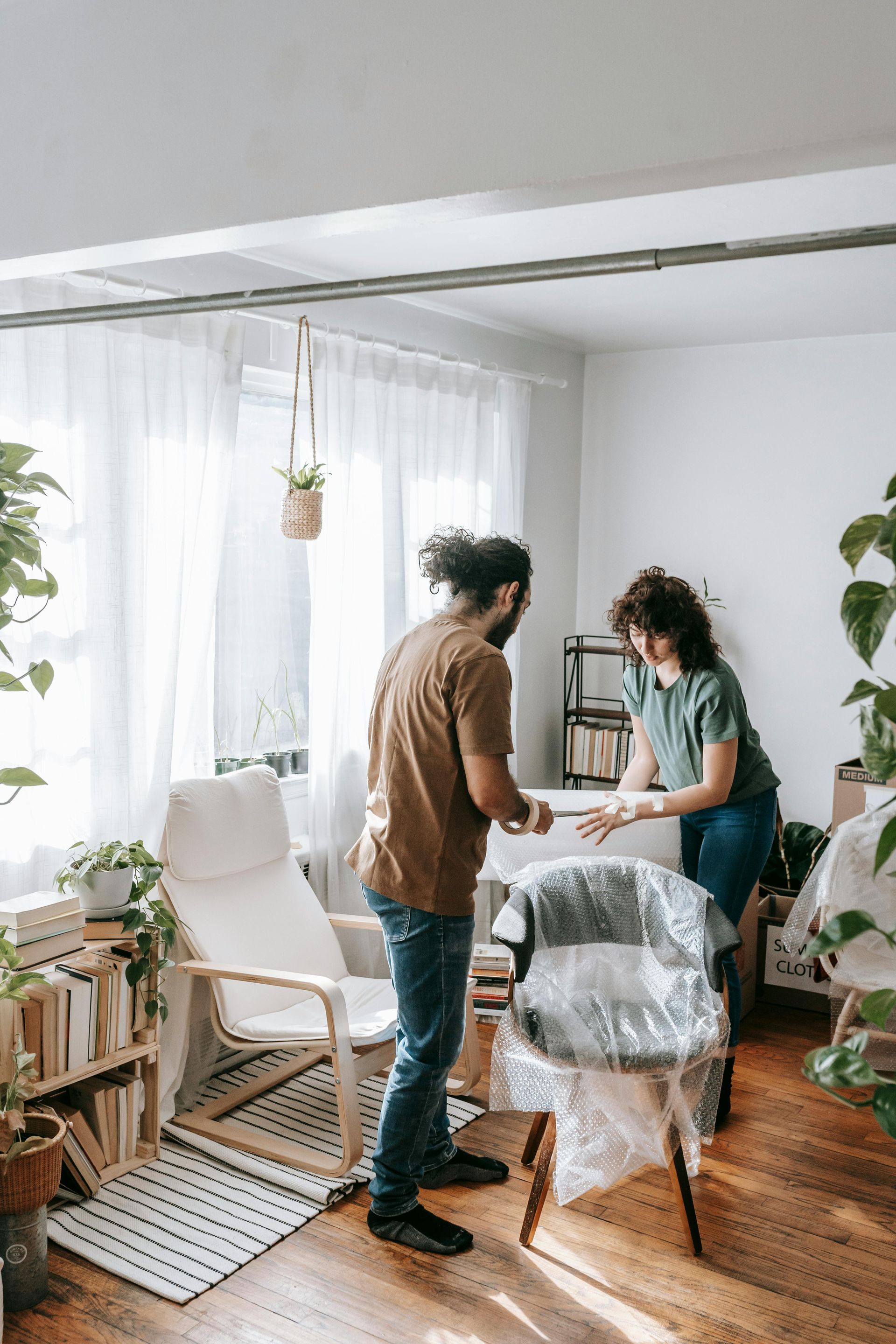 Couple unpacking furniture in a bright room with plants, wooden floors, and a sunny window.