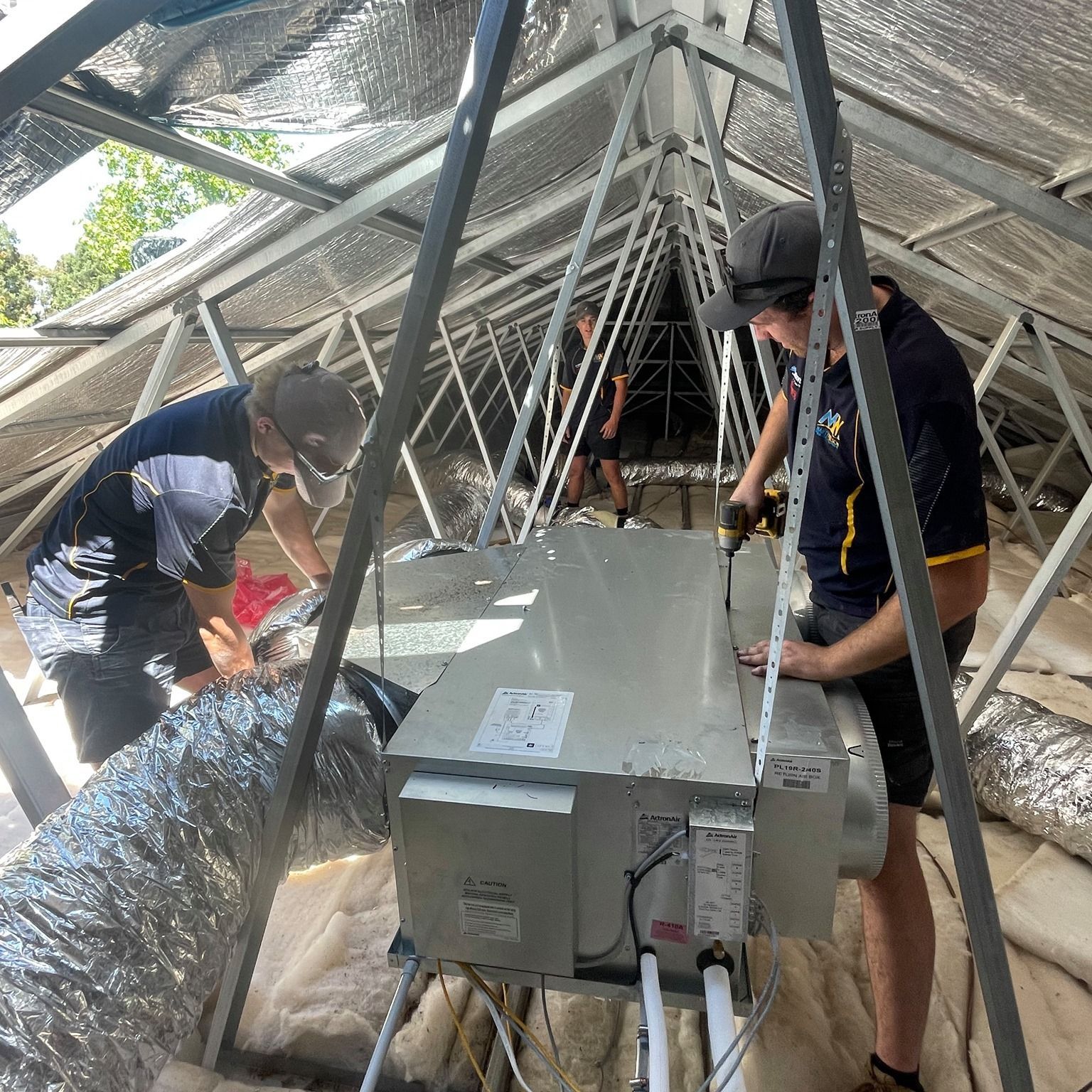 Technicians installing a ducted air conditioning system in a roof cavity, working with tools.