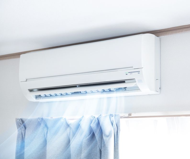 White wall-mounted air conditioner blowing cool air onto light blue curtains in a room.