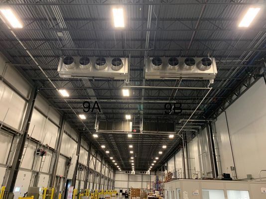Interior view of a high-ceiling industrial warehouse showing overhead lighting and two large, suspended refrigeration or air handling units labeled 