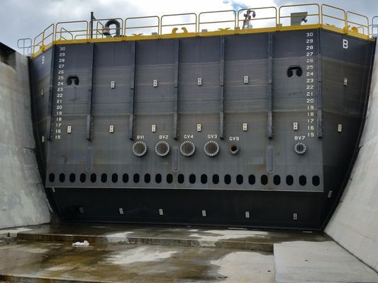 A massive, dark grey industrial metal gate with circular portholes and measurement scales.