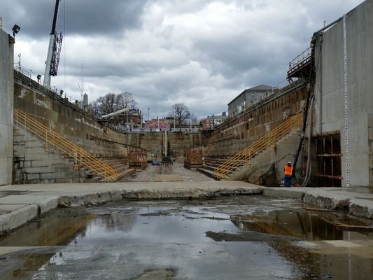 An empty concrete dry dock with workers and scaffolding under a cloudy sky.