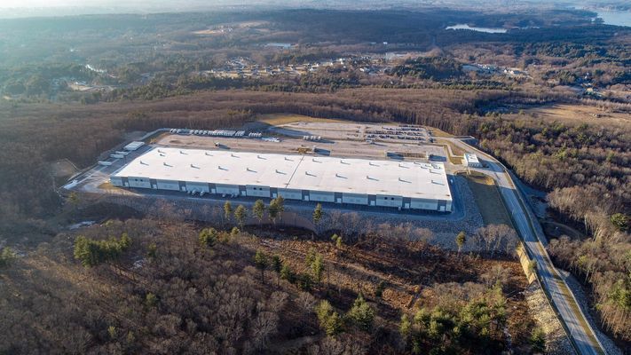 A wide aerial shot of a long white warehouse surrounded by dense forest.