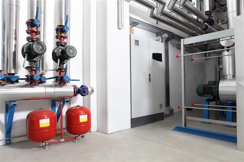 Two bright red vertical expansion tanks mounted below a network of insulated silver pipes and dual circulation pumps in a clean industrial facility.