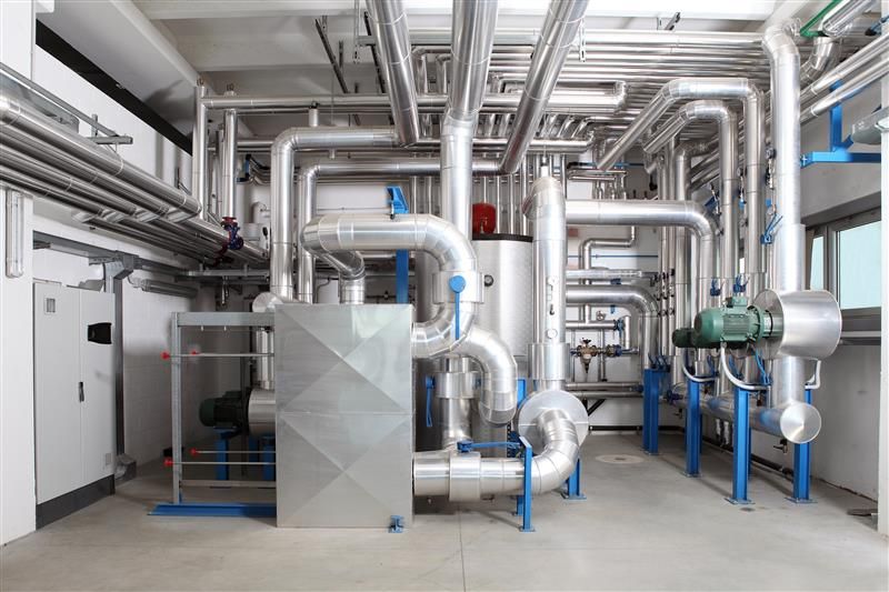 An intricate maze of silver-insulated industrial pipes and vents filling a large mechanical room, supported by blue metal frames.
