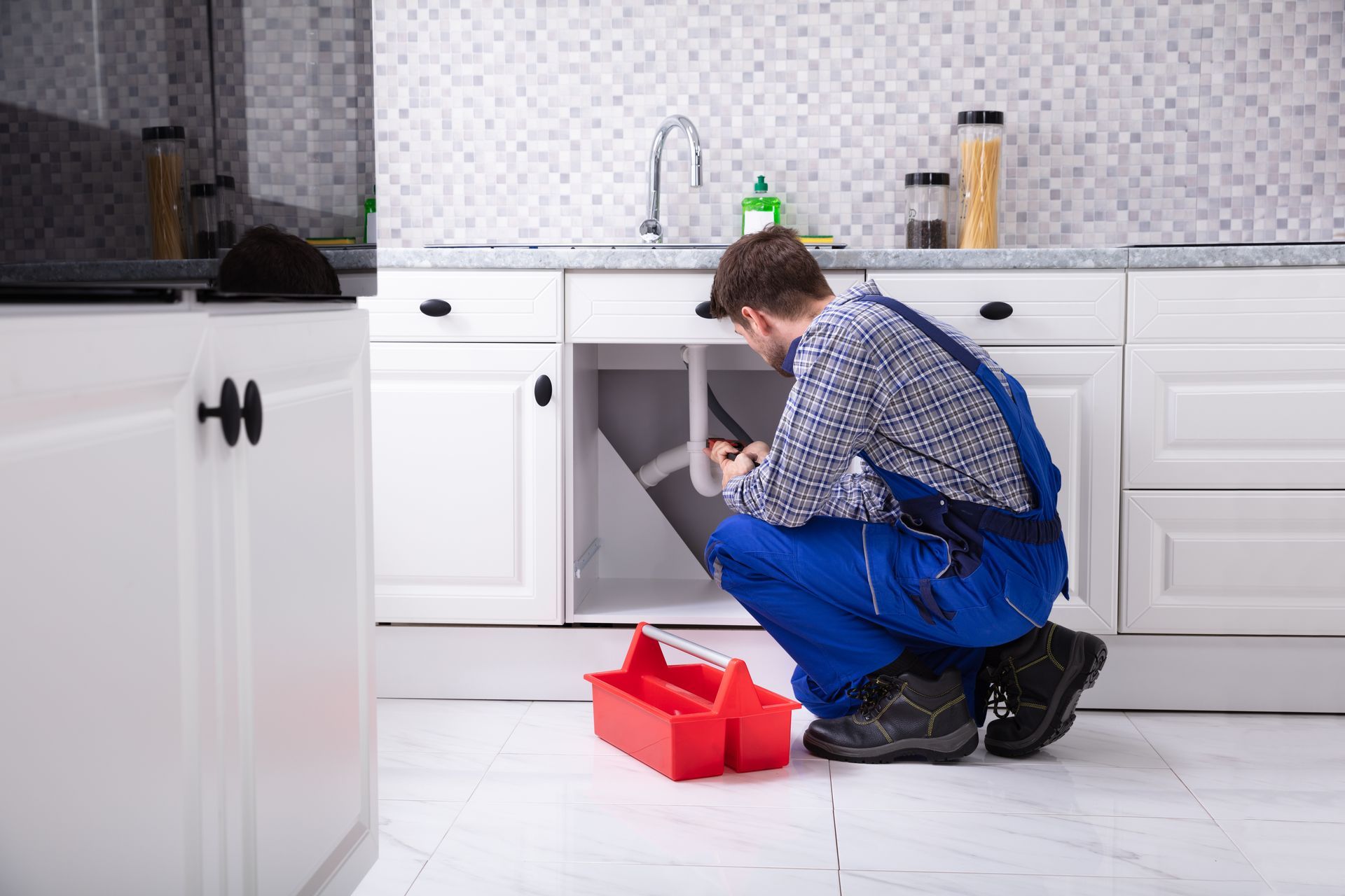 A plumber in blue overalls kneels on a white tile floor to inspect the drainage pipes under a kitchen sink, with a red toolbox nearby.