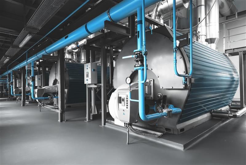 A line of heavy-duty horizontal industrial boilers with metallic blue cladding and silver accents installed along a grey floor in a factory.