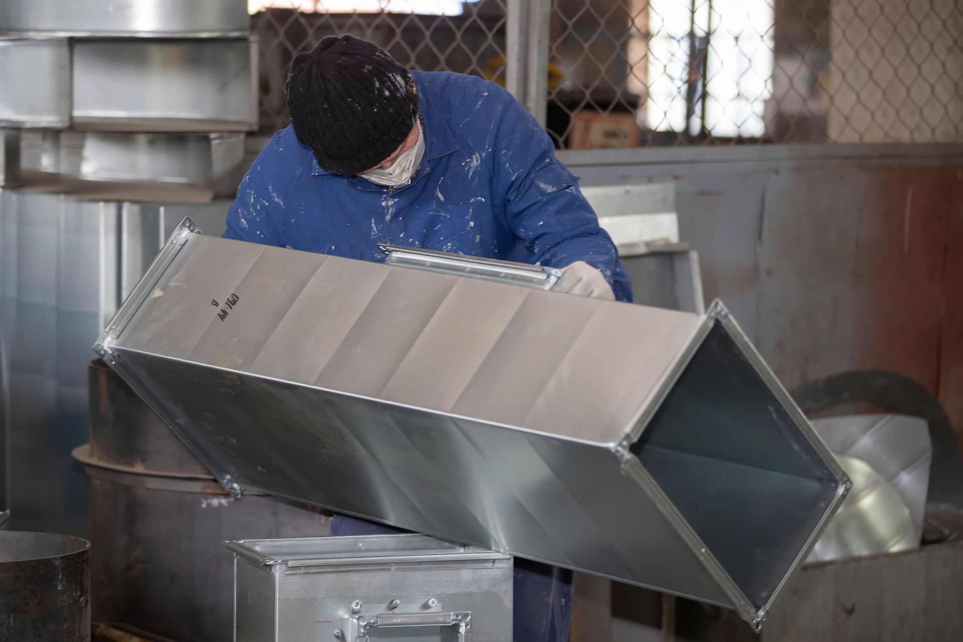 Sheet metal technician inspecting a rectangular duct section in a fabrication workshop.