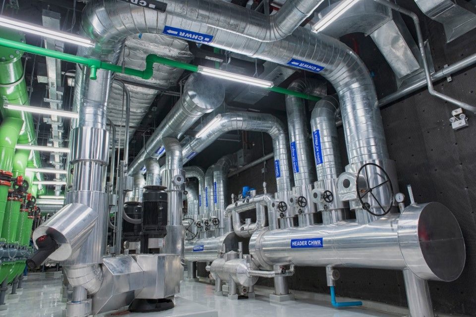 High-angle shot of a mechanical plant room featuring thick silver-wrapped pipes labeled with flow direction and contrasting green industrial gas lines.