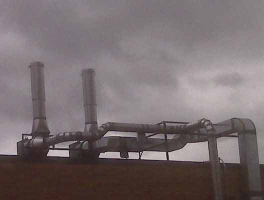 A series of horizontal and vertical silver exhaust vents and ductwork installed on a rooftop under a cloudy sky.