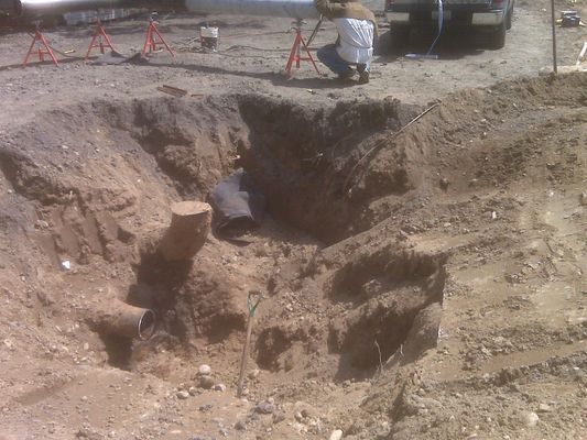 A wide excavation hole in the ground showing underground utility pipes and exposed soil.