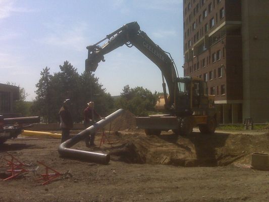 A yellow excavator lifting a large curved section of metal piping at an outdoor construction project.