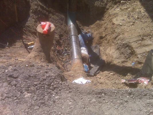 A top-down view of a large steel pipe joint being fitted within a narrow dirt trench.