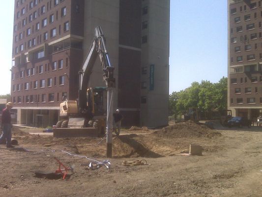 A yellow excavator with a hydraulic attachment working on a dirt construction site in front of a multi-story brick building.