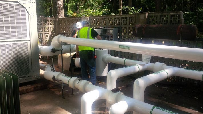 A technician in a high-visibility vest inspecting a network of large white insulated pipes at an outdoor mechanical installation.
