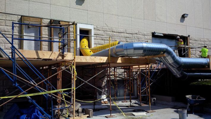 Exterior construction showing large metal ductwork entering a building through a temporary scaffolding setup.