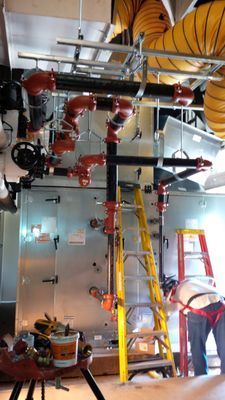 A technician on a ladder performing maintenance on overhead red industrial pipes and mechanical equipment.