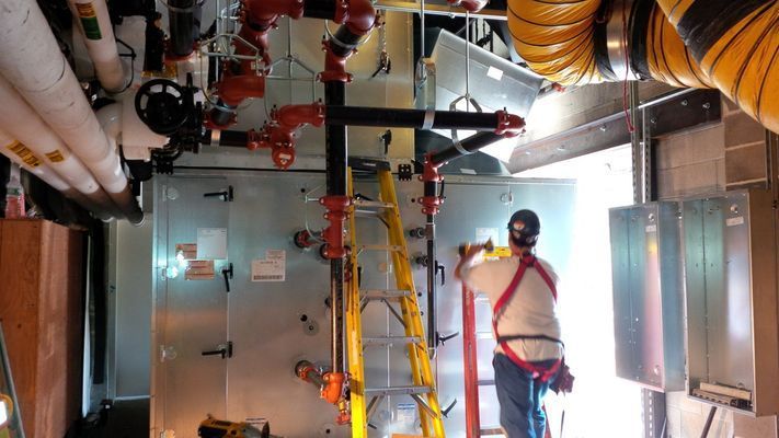 A technician in a safety harness working on the interior piping and electrical connections of a large commercial air handling unit.
