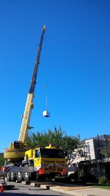 The yellow crane boom positioned over a residential or commercial area, preparing to lower a unit onto a roof.