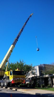 A yellow mobile crane extending its boom to lift a heavy HVAC component into the air against a clear blue sky.