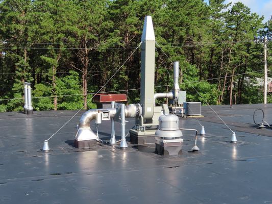 Rooftop exhaust system featuring a tall silver ventilation stack and associated ductwork against a backdrop of green trees.
