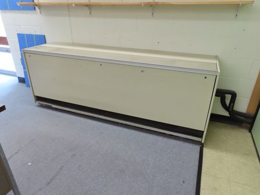 A cream-colored metal enclosure for a floor-mounted terminal heating or cooling unit installed along a white wall.
